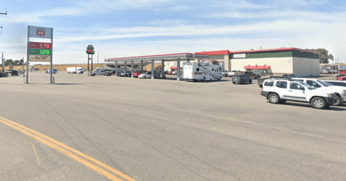 Gas station with multiple pumps, convenience store, and parked vehicles under a clear blue sky.