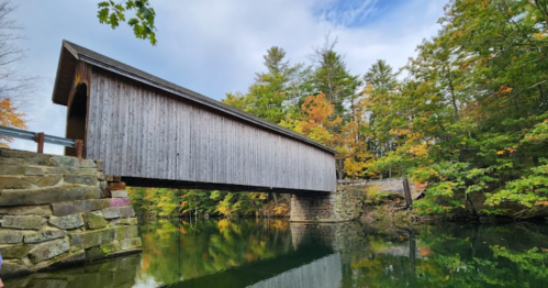 A wooden covered bridge spans a calm river, surrounded by colorful autumn trees and a cloudy sky.