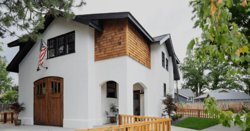 A modern two-story house with a wooden door, white exterior, and a small porch, surrounded by greenery.