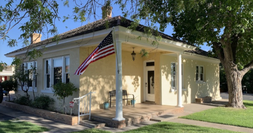 A yellow house with a porch, an American flag hanging, and a large tree nearby on a sunny day.