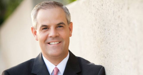 A smiling man in a suit stands against a textured wall, exuding confidence and approachability.