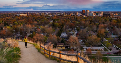 A person walks along a path overlooking a cityscape with trees and buildings under a colorful sky.