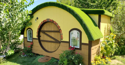 A whimsical yellow hobbit-style house with a round door, green grass roof, and surrounded by colorful flowers.