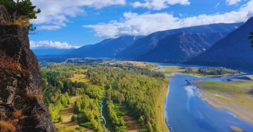 A scenic view of a river winding through lush green valleys and mountains under a bright blue sky.