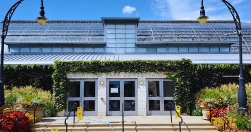 A modern conservatory building with glass panels, surrounded by greenery and colorful flowers under a blue sky.
