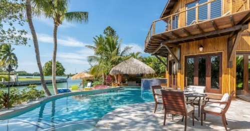 Tropical poolside view of a wooden house with palm trees, a thatched roof, and lounge chairs by the water.