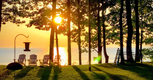 Sunset over a lake, with silhouetted trees and chairs on the shore, creating a serene outdoor scene.