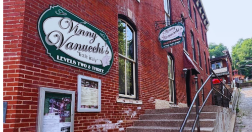 Exterior of Vinny Vanucci's Little Italy, featuring brick walls, signs, and a staircase leading to the entrance.