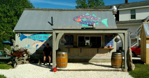 A colorful fish-themed food stand with a rustic exterior, featuring barrels and a menu board outside.