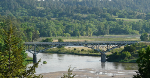 A metal bridge spans a river, surrounded by lush green trees and rolling hills in the background.