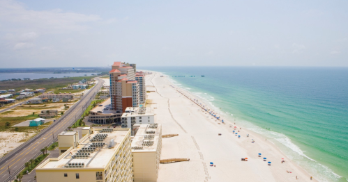 Aerial view of a sandy beach with turquoise water, lined by tall buildings and a road in the background.