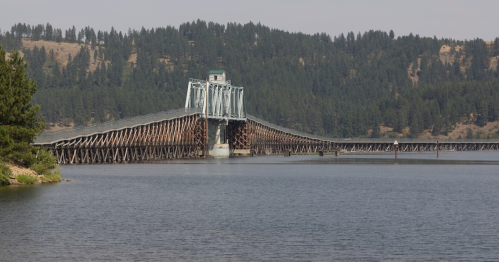 A long wooden bridge extends over a calm lake, surrounded by green hills and trees under a clear sky.