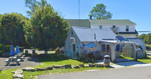 A colorful, rustic building with outdoor seating and picnic tables, surrounded by greenery and blue skies.