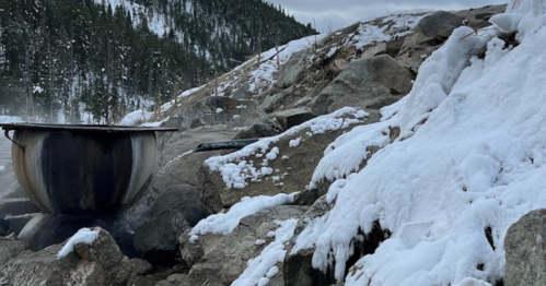 A large pot sits on rocky terrain, surrounded by snow and mist, with a forested mountain in the background.