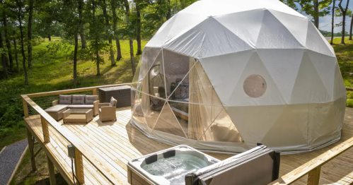 A geodesic dome on a wooden deck, featuring outdoor seating and a hot tub, surrounded by trees.