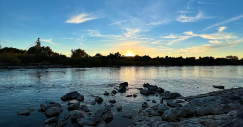 Sunset over a calm river, with rocky shorelines and trees silhouetted against a colorful sky.