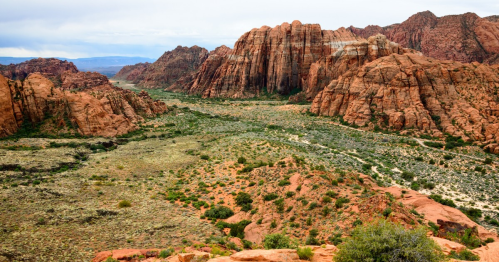 A panoramic view of rugged red rock formations and a lush green valley under a cloudy sky.