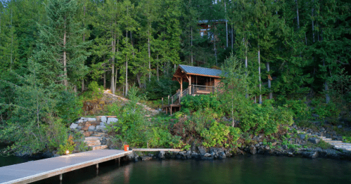 A wooden cabin nestled among lush green trees by a calm lake, with a wooden dock extending into the water.