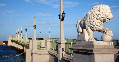 A lion statue stands beside a green bridge with lampposts, overlooking a calm body of water under a blue sky.