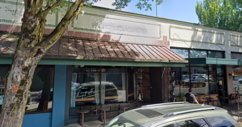 A street view of a storefront with a brown awning, trees nearby, and people walking by.