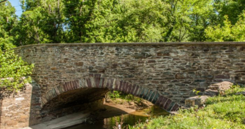 A stone arch bridge over a shallow creek, surrounded by lush green trees and foliage.
