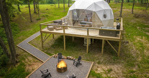 A geodesic dome cabin on a wooden deck, surrounded by trees and a fire pit with seating in a grassy area.