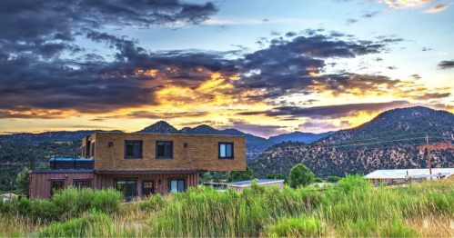 A modern house surrounded by greenery, set against a vibrant sunset and mountain backdrop.