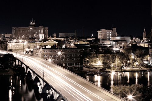 A nighttime cityscape featuring a bridge, illuminated buildings, and reflections on the water.