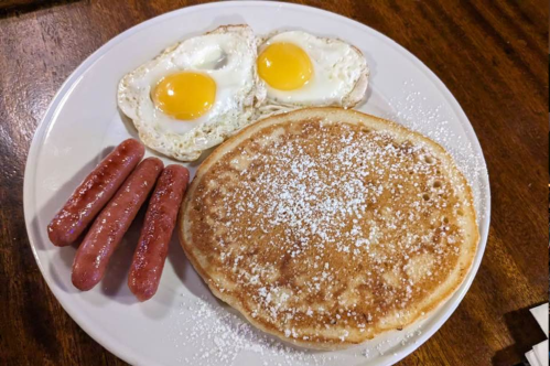 A plate with two fried eggs, three sausages, and a large pancake dusted with powdered sugar.