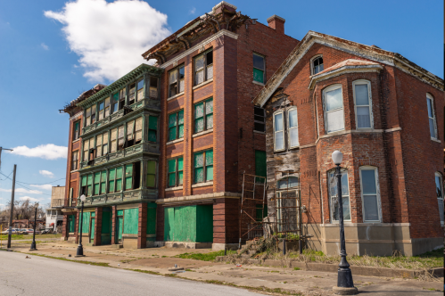 Abandoned brick buildings with boarded windows and peeling paint under a blue sky.