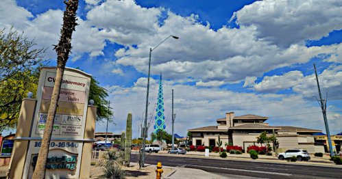 A street view featuring a pharmacy sign, a tall decorative tower, and a cloudy blue sky in a desert setting.