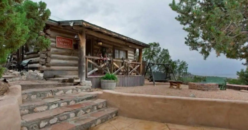 A rustic log cabin with a porch, surrounded by trees and a stone pathway, set against a cloudy sky.
