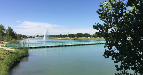 A serene lake with a fountain, surrounded by greenery and a walkway, under a clear blue sky.