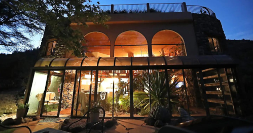 A unique stone and glass house at dusk, featuring arched windows and a lush garden.