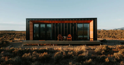 A modern cabin with large windows and wooden accents, set in a desert landscape with sparse vegetation.