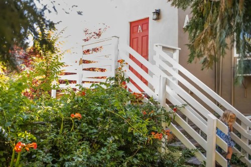 A child walks down white stairs surrounded by greenery and orange flowers, leading to a red door.