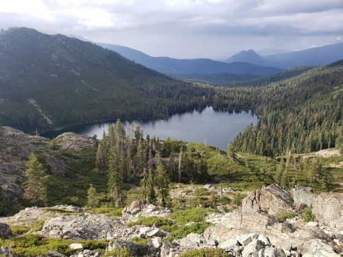 A scenic view of a tranquil lake surrounded by mountains and dense forests under a cloudy sky.