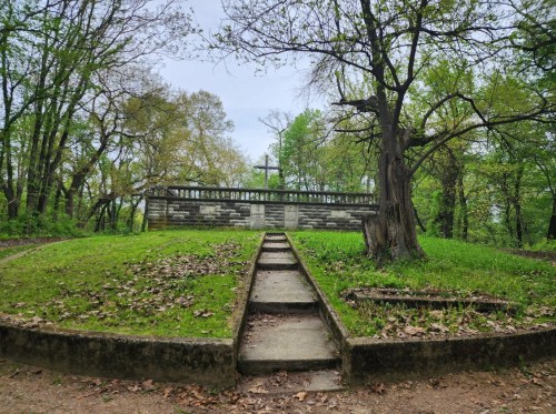 A stone memorial surrounded by trees, with a cross and steps leading up to it, set in a green, grassy area.