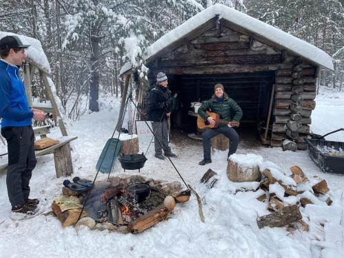 Two people near a log cabin in the snow, one playing guitar by a campfire, another cooking. Snow-covered trees in the background.