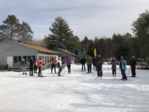 A group of people in winter gear stand on snow, ready for cross-country skiing near a wooden building and trees.