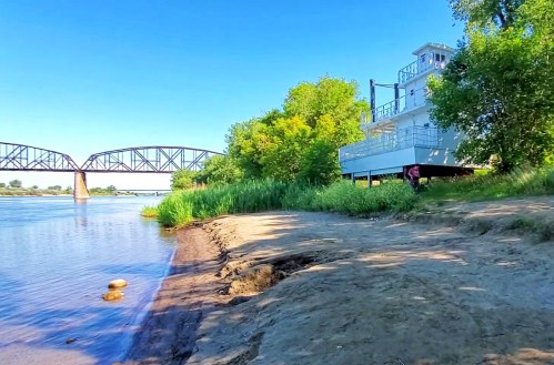 A sandy riverbank with lush greenery, a boat docked nearby, and a bridge in the background under a clear blue sky.