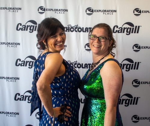 Two women in elegant dresses pose playfully at a photo backdrop featuring logos for Exploration Place and Cargill.