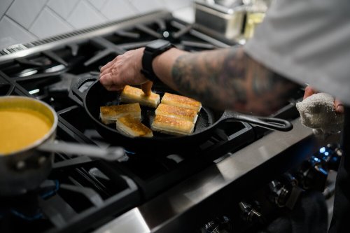 A chef's hand places golden-brown slices in a cast iron skillet on a stovetop, with a pot of soup nearby.