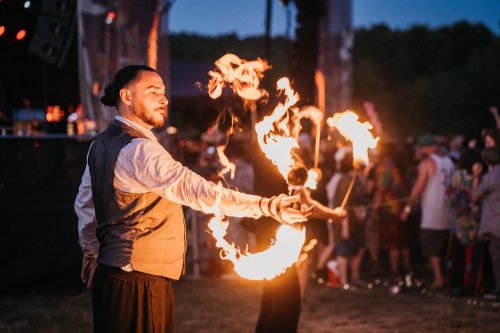 A fire performer twirls flaming torches at a festival, with a crowd enjoying the show in the background.