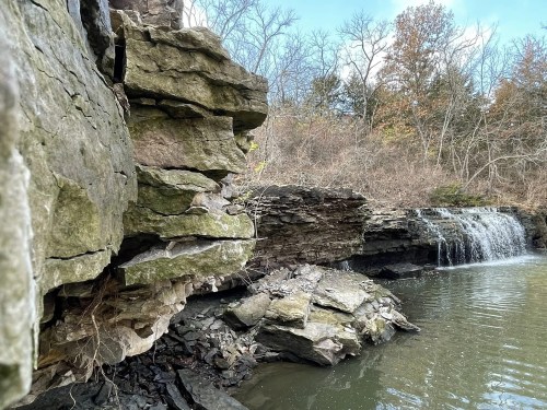 Rocky cliffside beside a calm water body, with a small waterfall cascading into the water, surrounded by trees.
