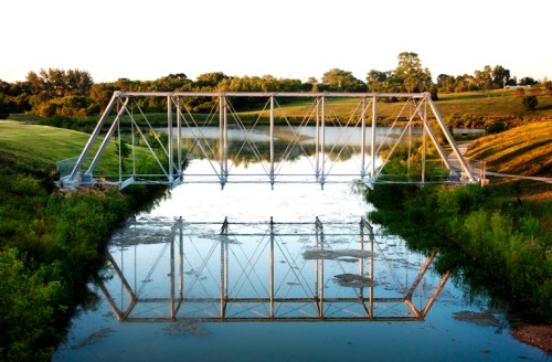 A metal bridge spans a calm river, reflecting its structure in the water, surrounded by lush greenery and trees.