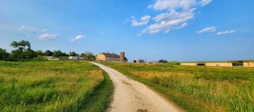 A gravel path leads through green fields to a farm with buildings under a blue sky with scattered clouds.