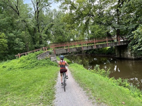 A person rides a bike along a path by a river, with a bridge and lush greenery in the background.