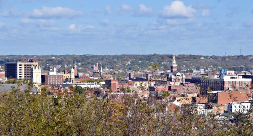 A panoramic view of a city skyline with various buildings and a church steeple, set against a blue sky with clouds.