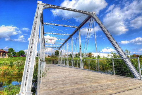 A metal truss bridge spans a calm river, surrounded by greenery and a bright blue sky with fluffy clouds.